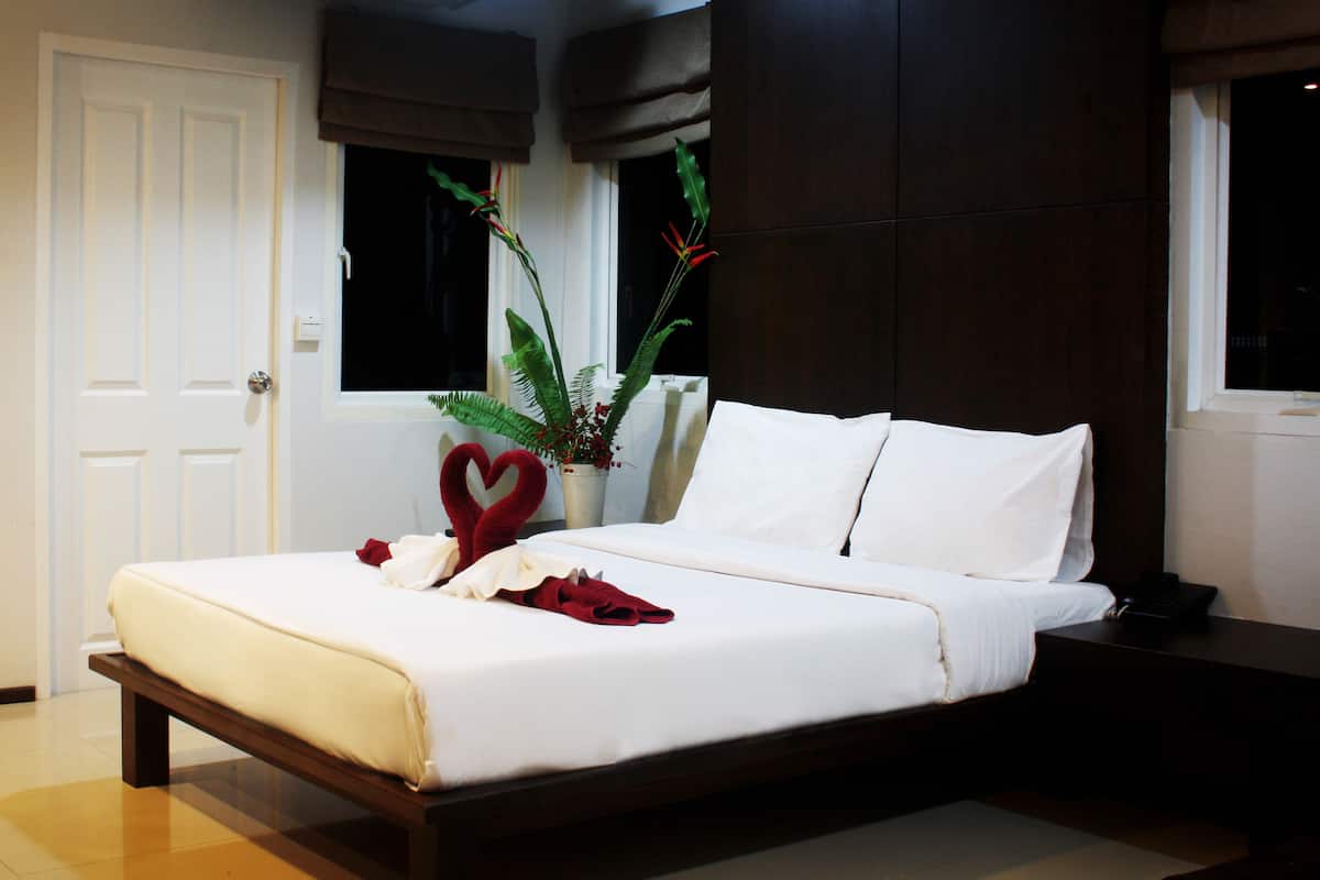 A neatly made bed with white linens, a heart-shaped decoration, and a vase with green plants.