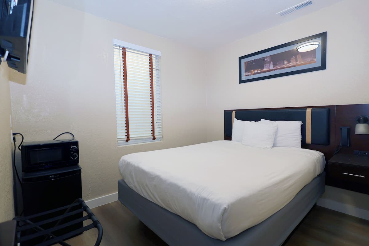 A hotel room with a bed, a nightstand, a lamp, a picture on the wall, a window with blinds, and a microwave on a cart.