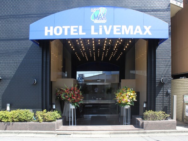 The entrance of Hotel Livemax with a blue awning and two flower arrangements.