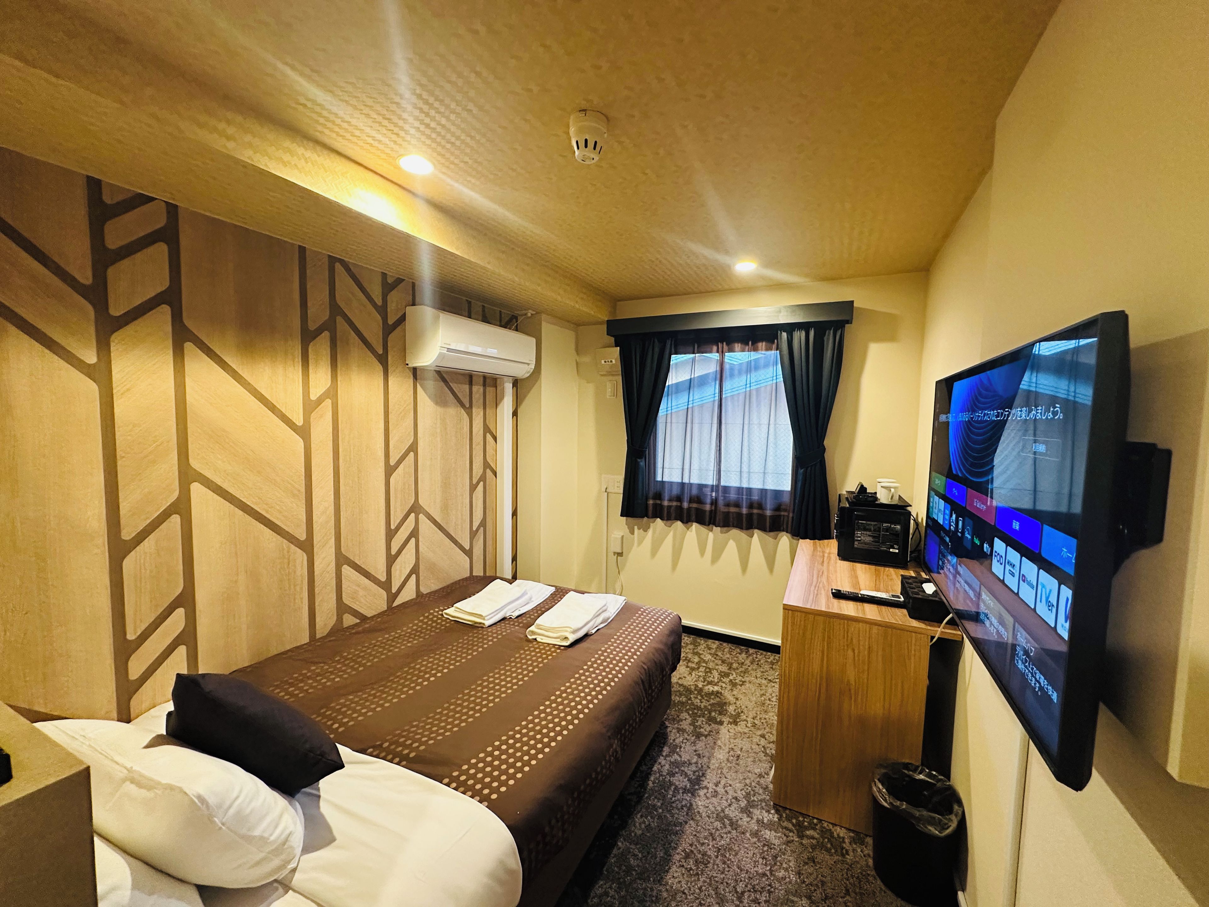 A hotel room with a bed, a desk with a television, an air conditioning unit, and a window with curtains.