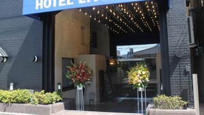 De ingang van Hotel Livemax met een blauw luifel, twee bloemstukken en een bakstenen gevel.