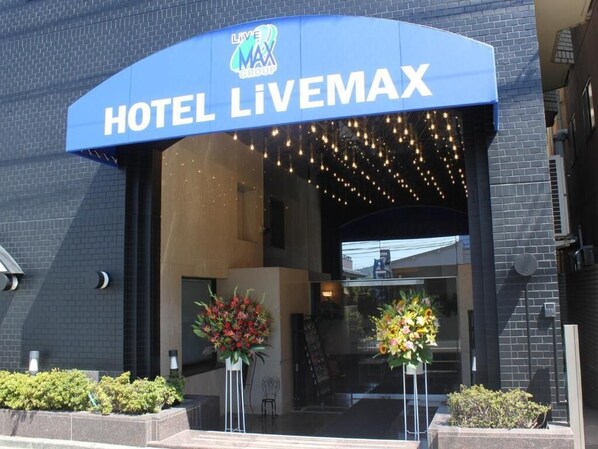 De ingang van Hotel Livemax met een blauw luifel, twee bloemstukken en een bakstenen gevel.