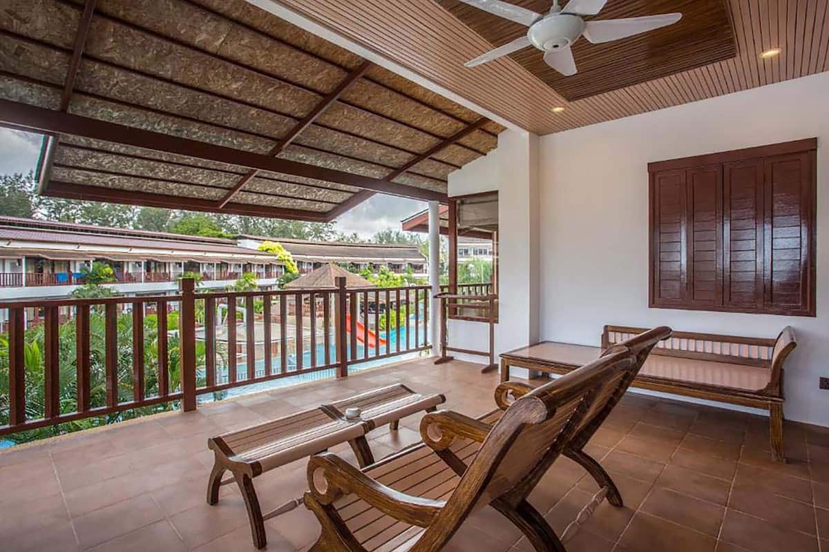 Área externa coberta com móveis de madeira, ventilador de teto e vista para uma piscina e edifícios ao fundo.