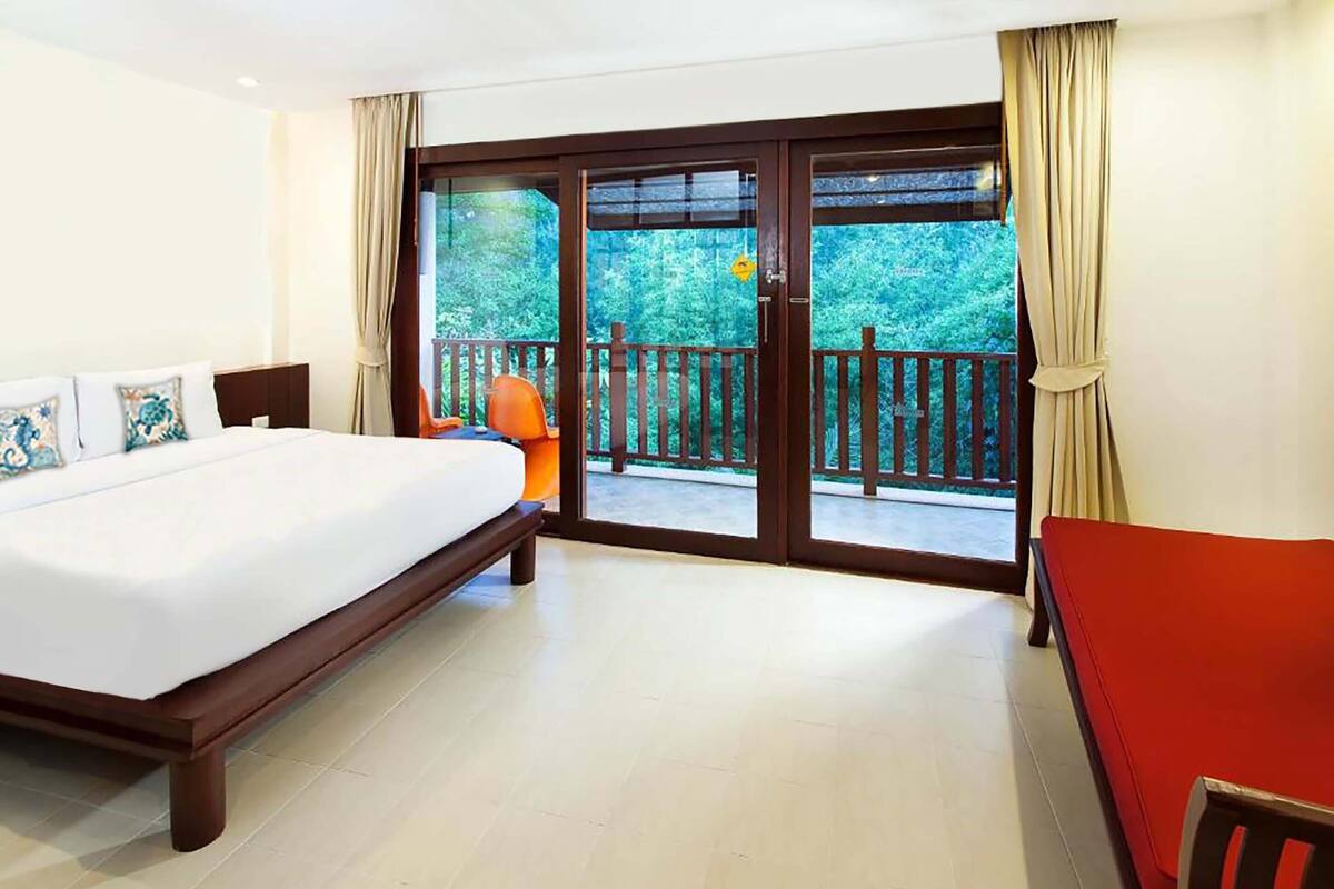 Quarto de hotel com uma cama, um sofá vermelho e uma varanda com grade de madeira.