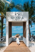 Un sendero de madera que lleva a un arco blanco con el nombre "ARINARA BEACH RESORT PHUKET" y palmeras al fondo.
