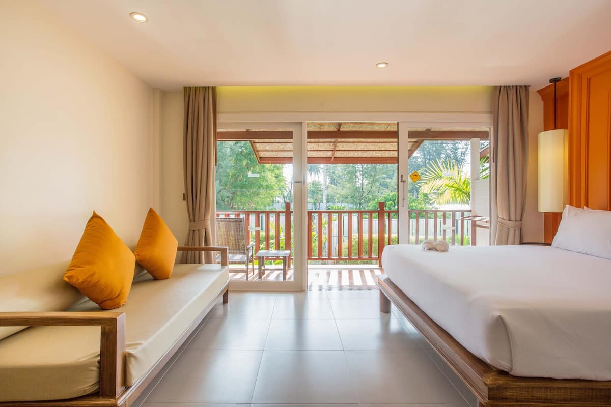 Quarto de hotel com cama, sofá, poltrona e varanda com vista para o verde.