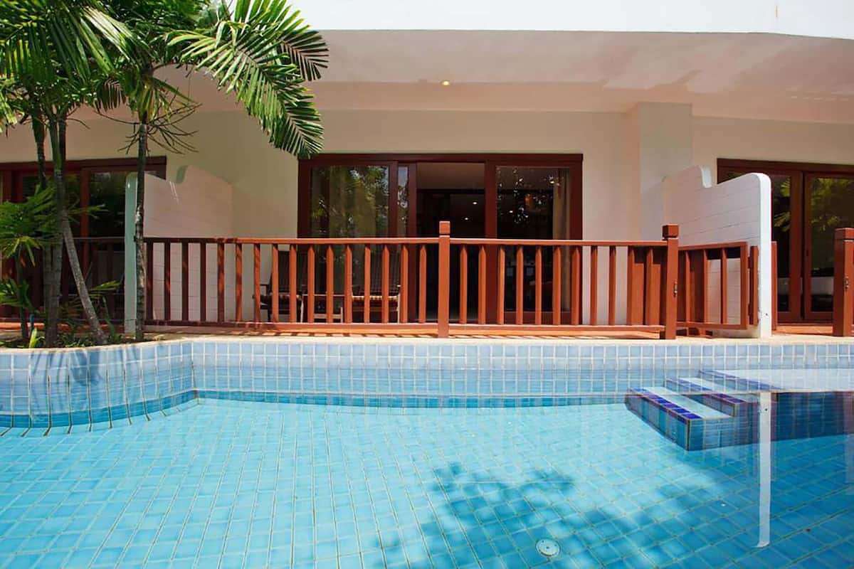 Piscina com deck de madeira e guarda-corpo de vidro, rodeada por palmeiras e um edifício com amplas janelas.