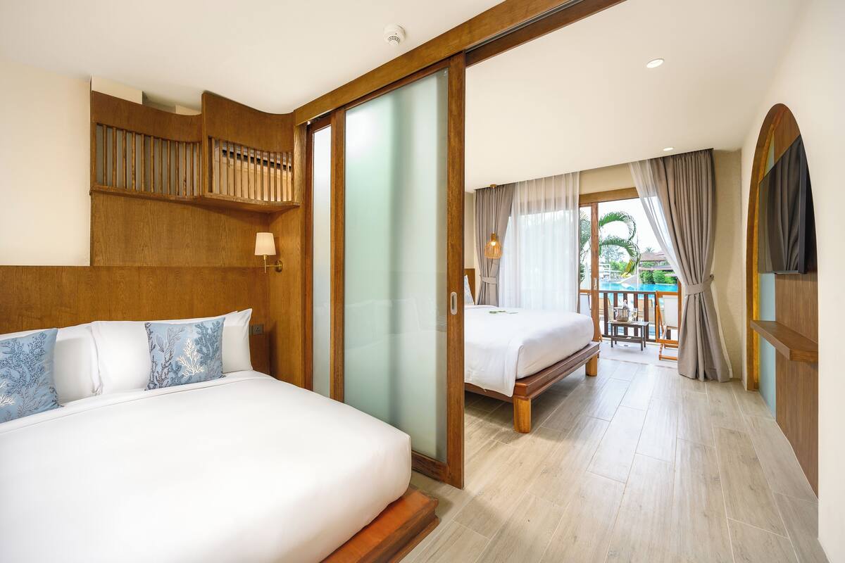 Quarto de hotel com uma cama grande, cabeceira de madeira, porta de vidro deslizante para uma varanda e vista para o exterior.