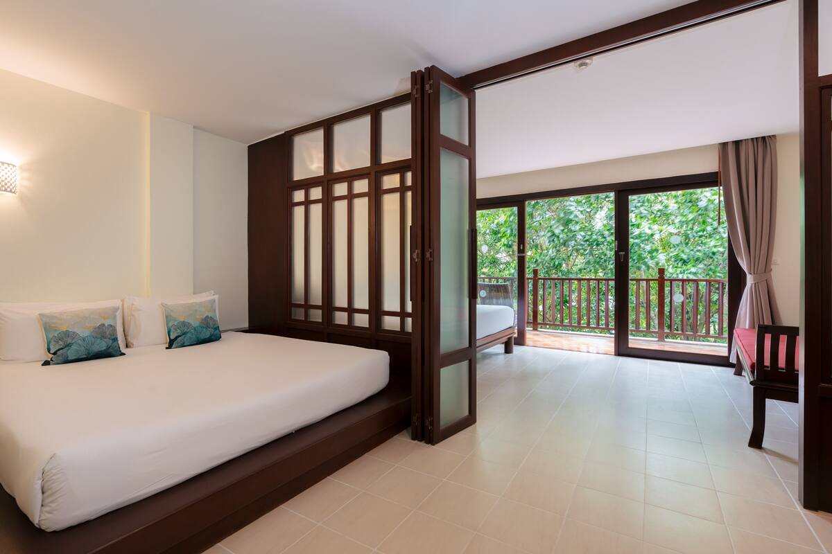Quarto de hotel com uma cama grande, porta de vidro deslizante para uma varanda e vista para o verde lá fora.