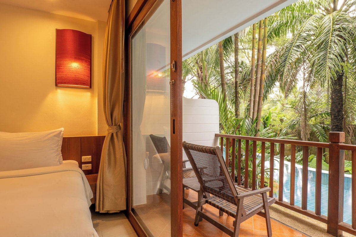 Habitación de hotel con cama, una silla, vista a una piscina y un balcón con barandilla.
