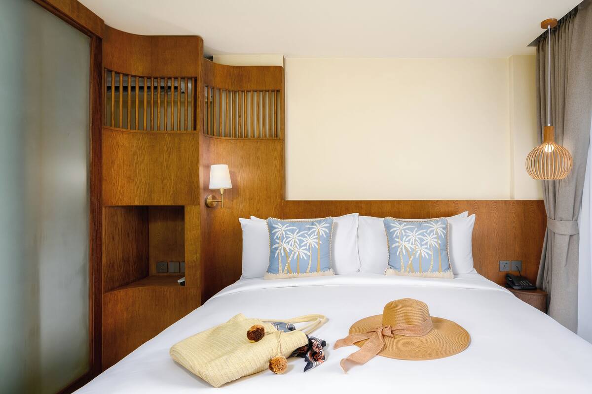 Quarto de hotel com cabeceira de madeira, cama com roupa de cama branca, duas almofadas decorativas, mesa de cabeceira com abajur e um chapéu sobre a cama.