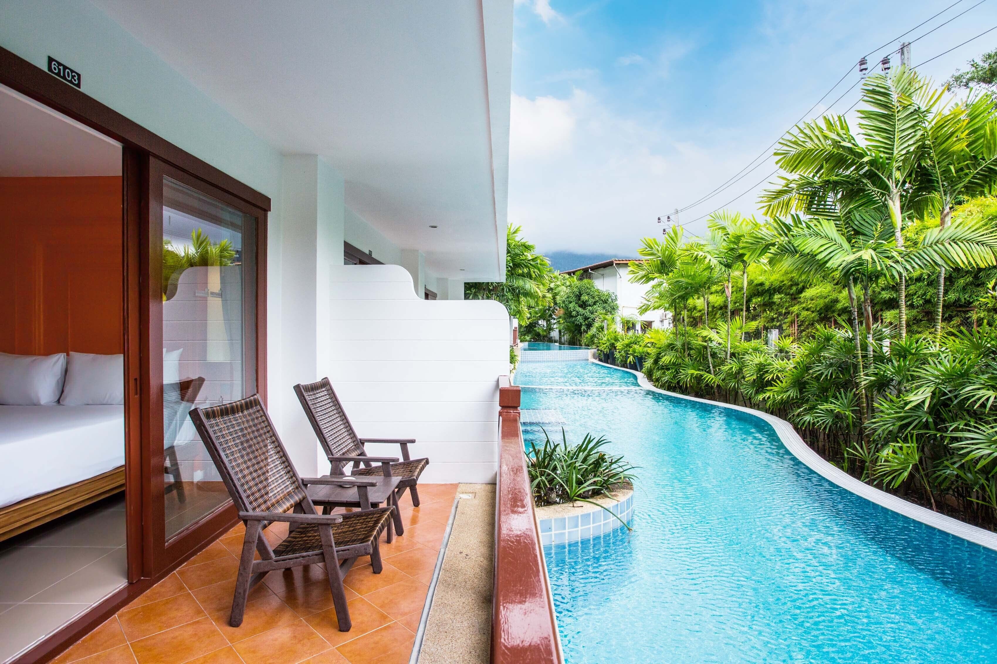 Quarto de hotel moderno com porta de vidro, um sofá, duas cadeiras de vime e vista para uma piscina curva cercada por vegetação exuberante.