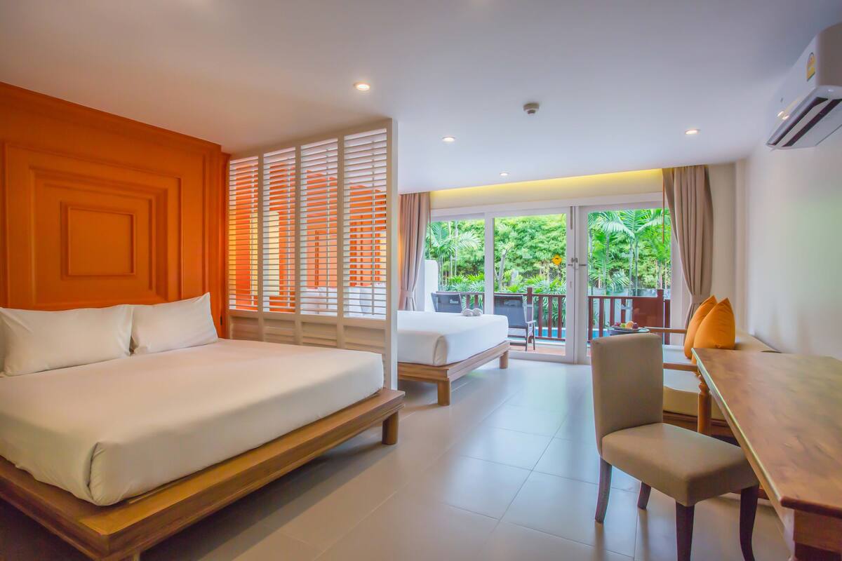 Quarto de hotel com uma cama grande, uma cama menor, uma mesa de jantar e uma cadeira. Possui uma varanda com vista para áreas verdes.