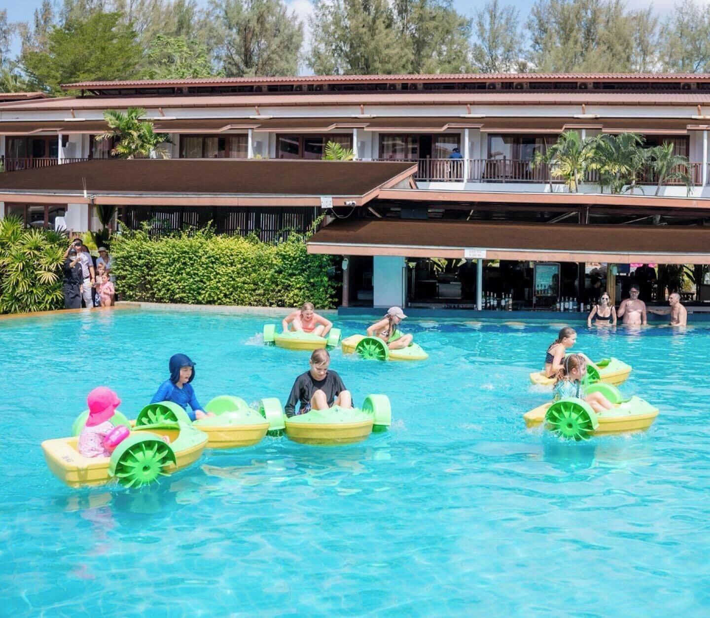 A swimming pool with people on watercrafts, a building with a roof and balconies, and greenery in the background.