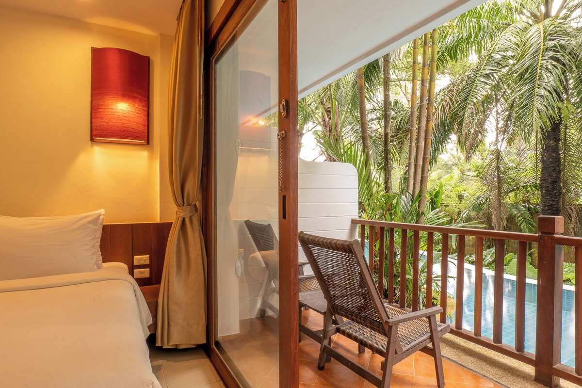 Habitación de hotel con cama, una silla, vista a una piscina y un balcón con barandilla.