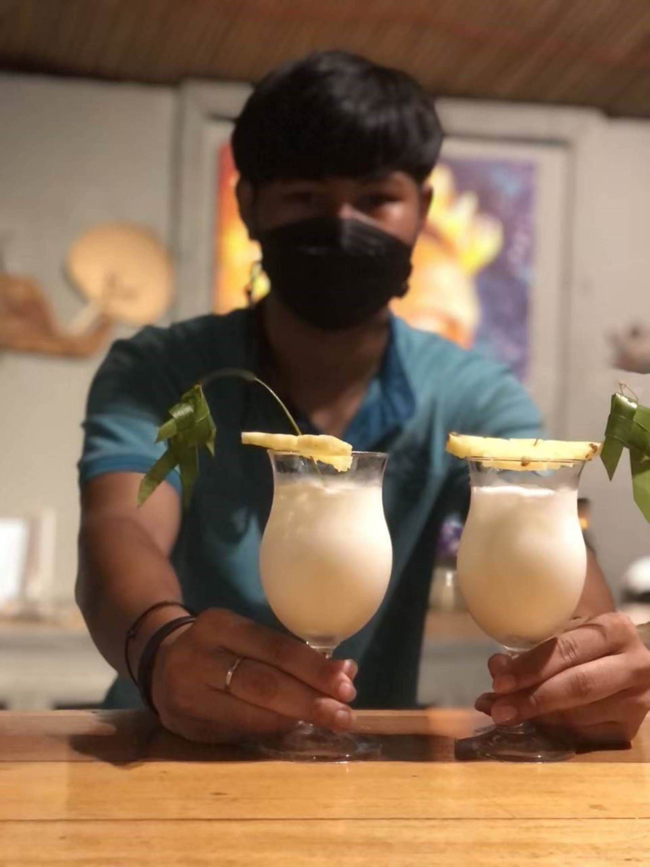 A person holding two glasses of a creamy beverage with a garnish, in a room with a wooden ceiling and a poster on the wall.