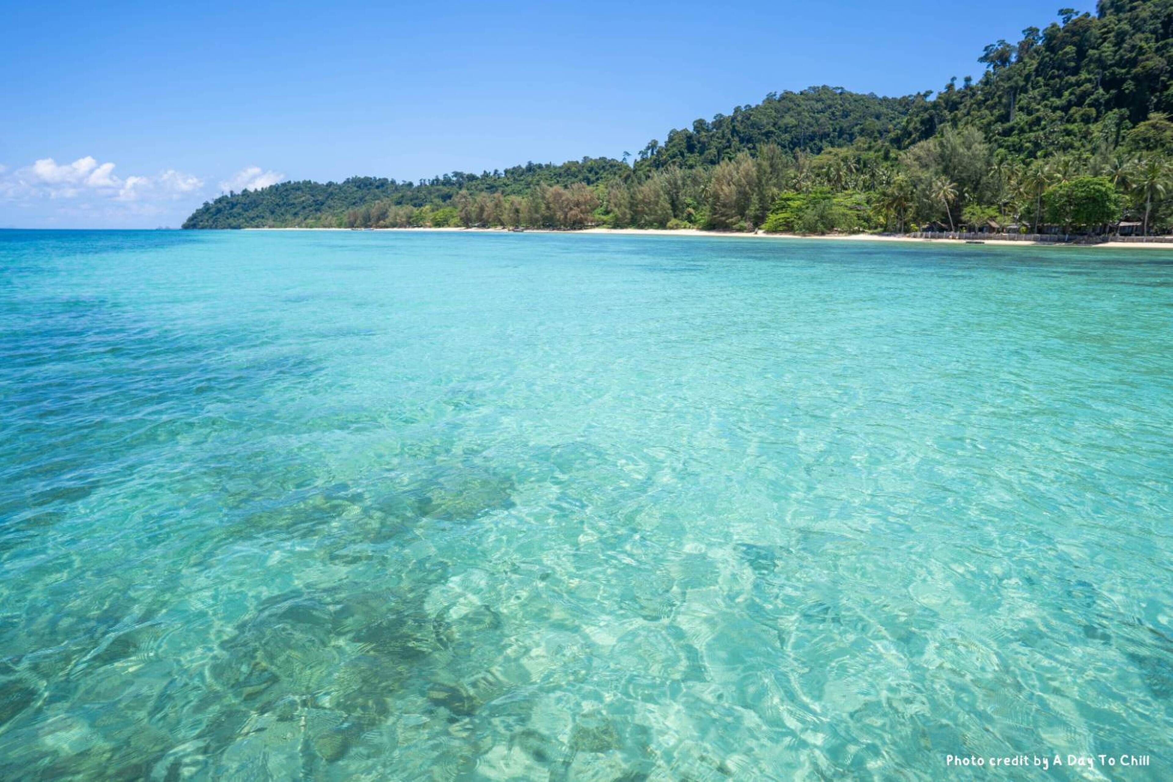 A clear turquoise sea with a sandy beach and a forested hill in the background.