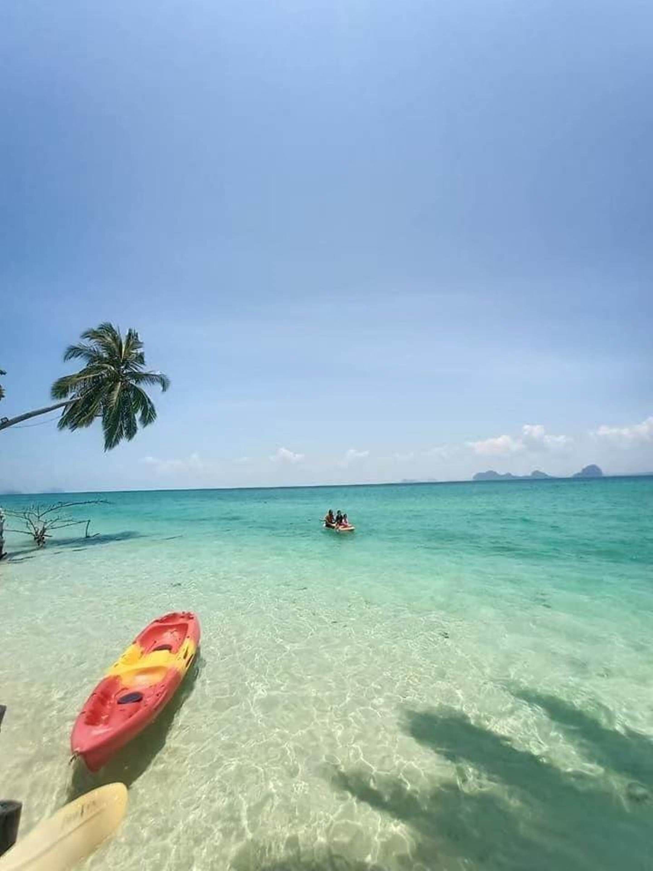 Kristalblauwe zee met een kajak en een persoon in het water, palmbomen en een heldere lucht.