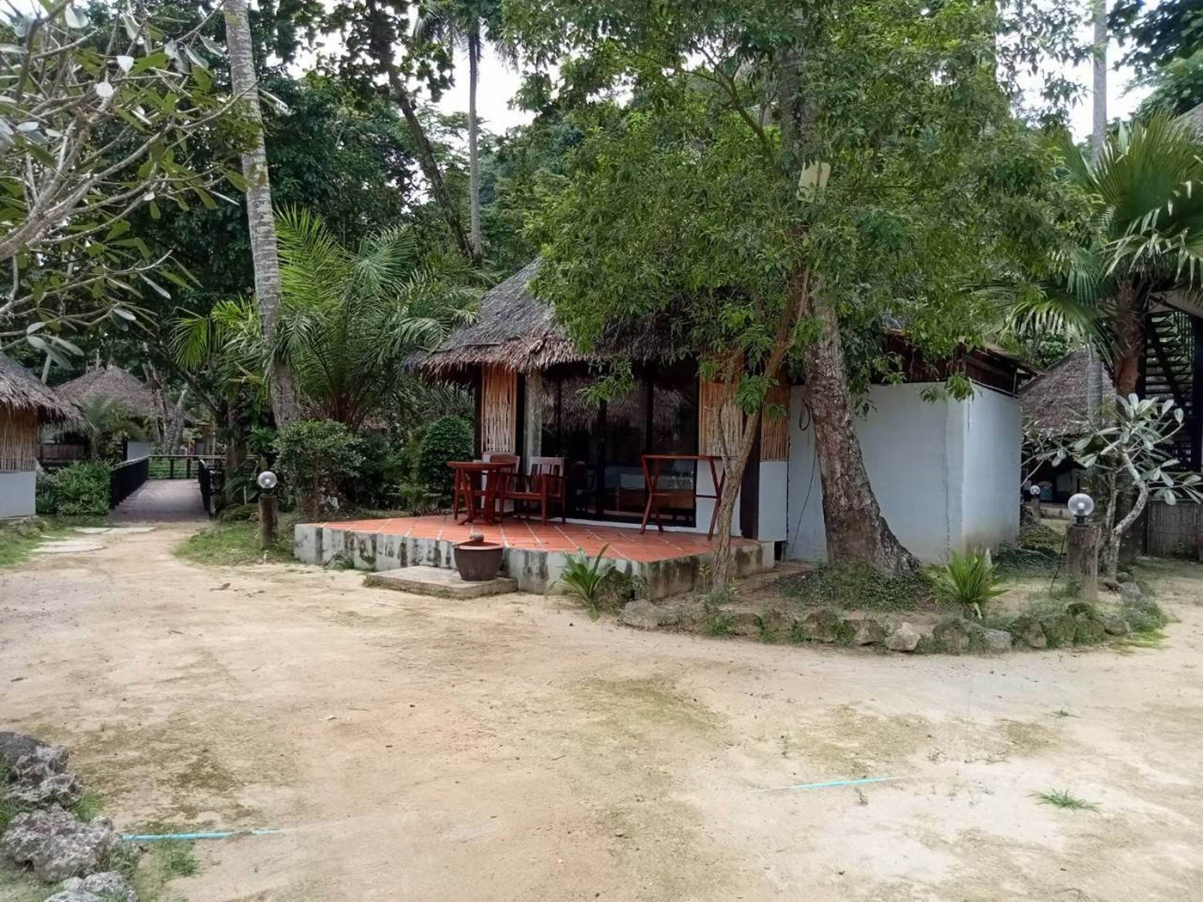 Un espacio de estar rústico al aire libre con una estructura de techo de paja, rodeado de abundante vegetación y árboles.