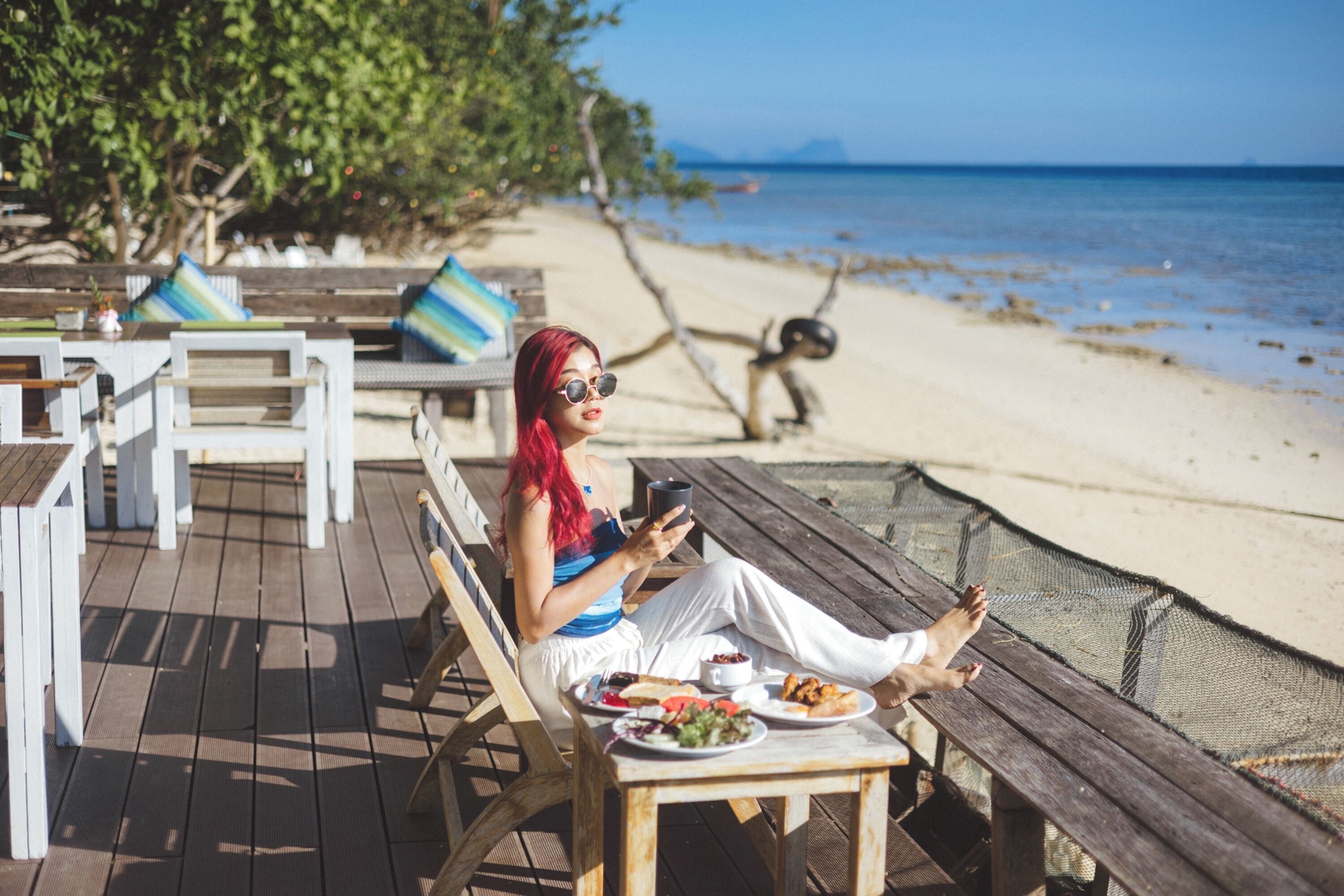 A person sitting on a wooden deck with a table of food and a cup, overlooking a beach.