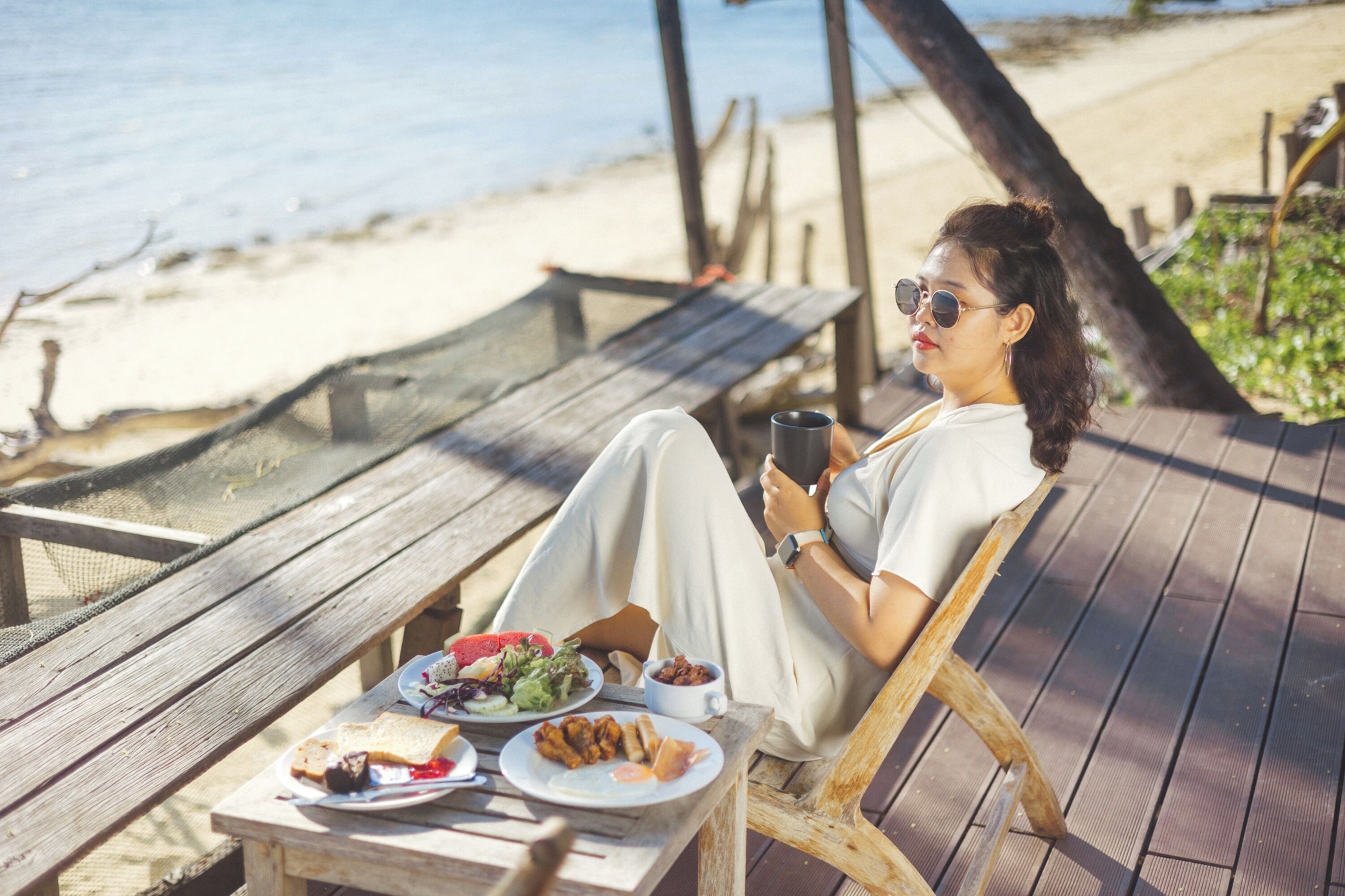 A person sitting on a wooden deck chair with a table in front of them, holding a cup.