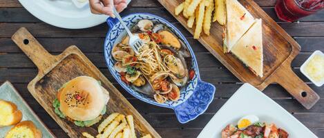 A variety of dishes on wooden serving boards, including a burger, fries, a seafood pasta, a sandwich, and a salad.