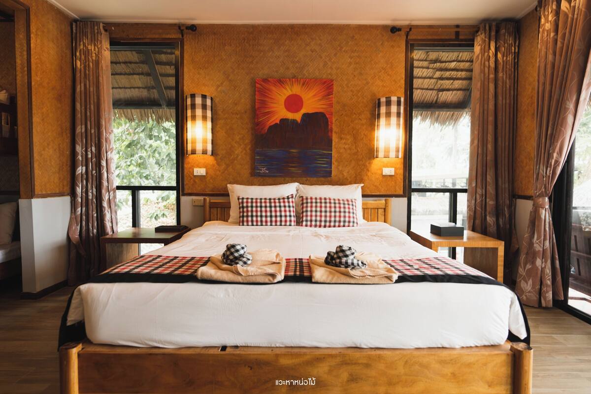 Una habitación de hotel con una cama grande, dos mesitas de noche, un cuadro de una puesta de sol y cortinas en las ventanas.