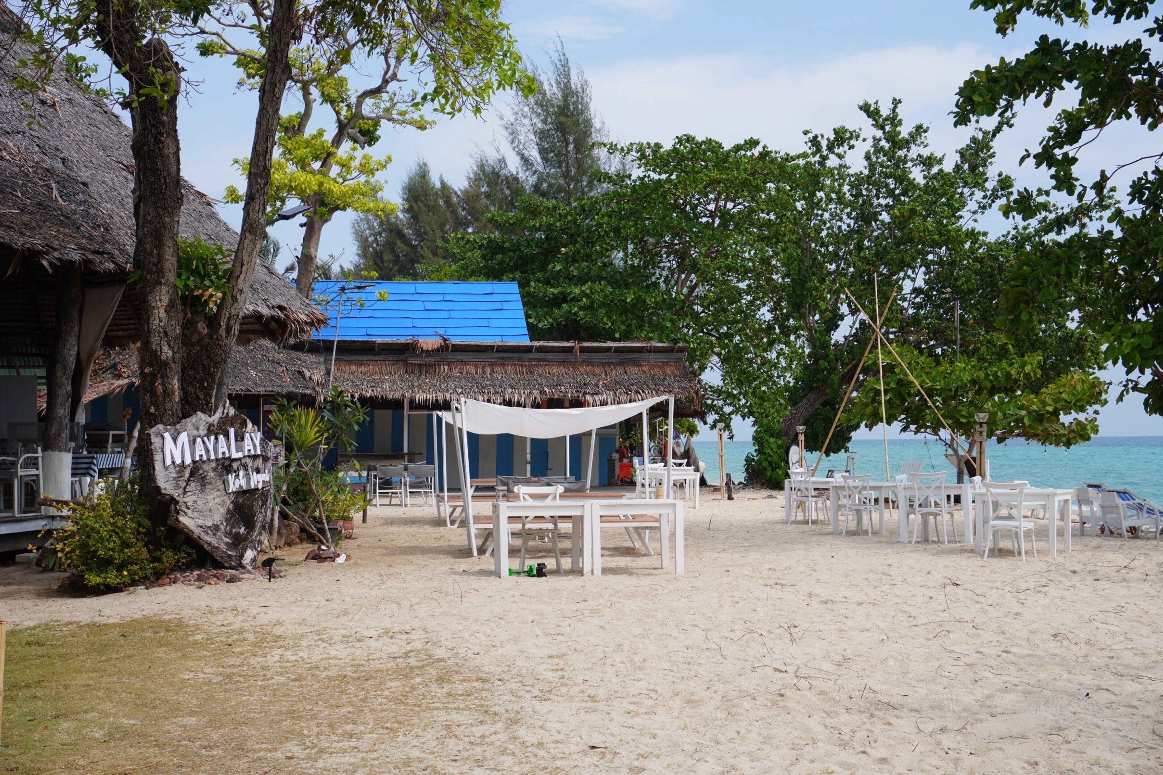 Un restaurant situé en bord de mer, doté d’une terrasse, d’un bâtiment à toit de chaume et d’une enseigne sur laquelle on peut lire « MAYALAY ».