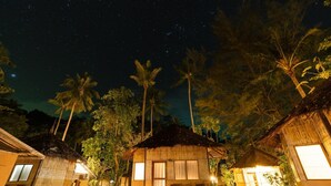Una cabaña con techo de paja, un pequeño porche y una mesa, rodeada de palmeras y otras cabañas bajo un cielo estrellado.