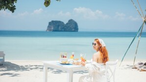 Una persona cenando en una mesa blanca en la playa, con vista al mar y una gran formación rocosa a lo lejos.