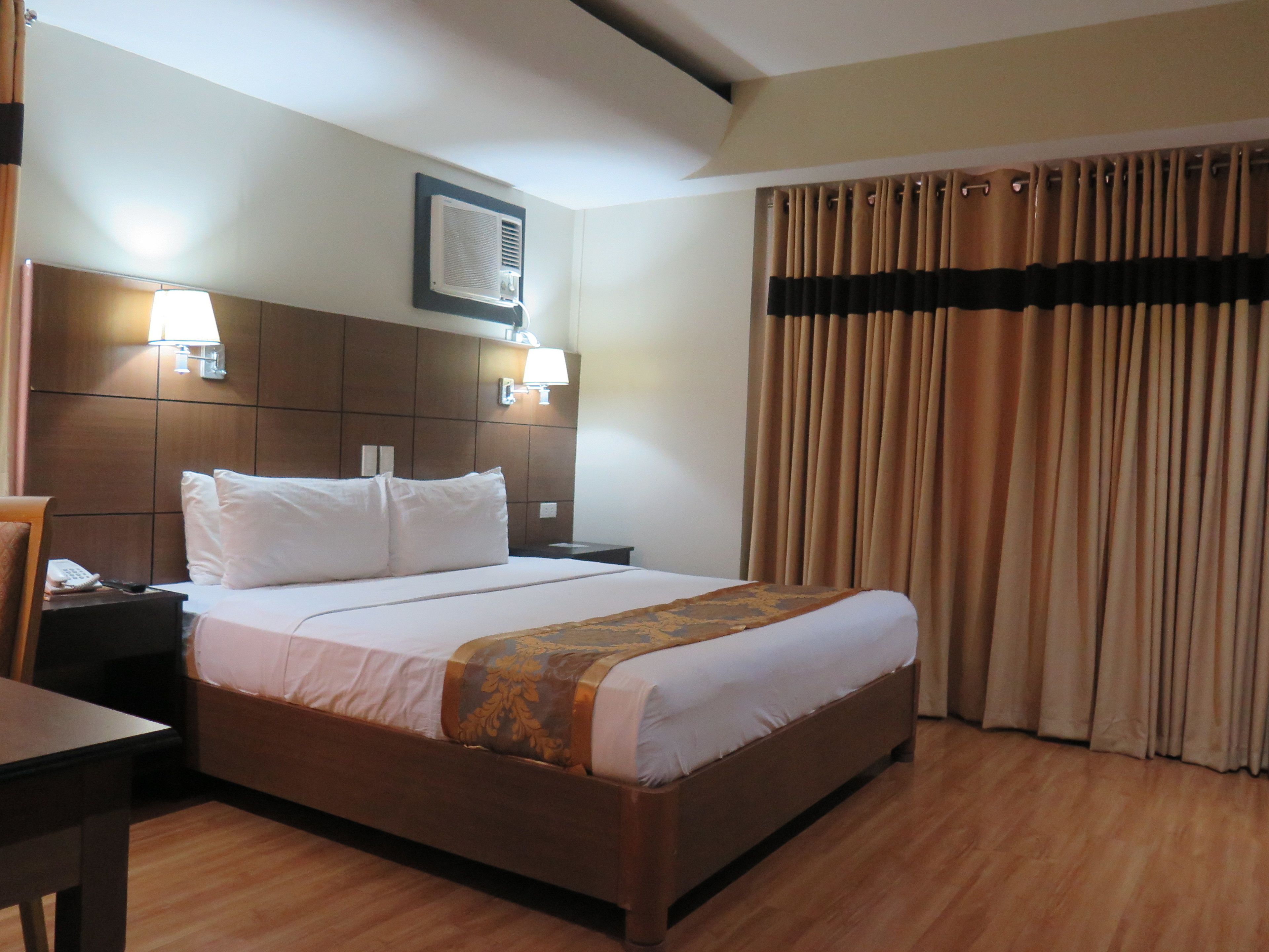 A hotel room with a wooden bed, a nightstand, a lamp, an air conditioning unit, and a curtain.
