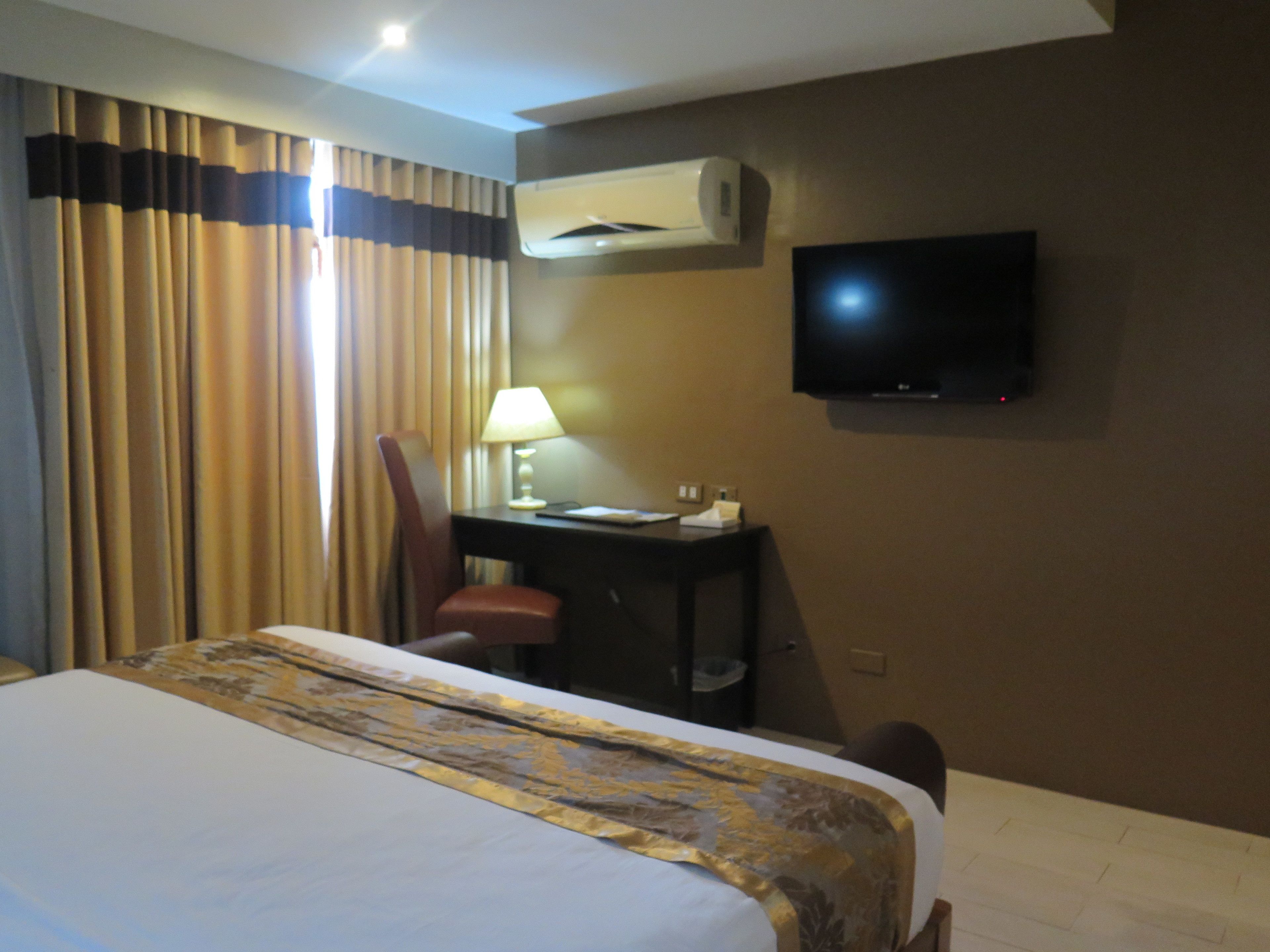 A hotel room with a bed, a desk with a lamp, a chair, an air conditioning unit, and a television mounted on the wall.
