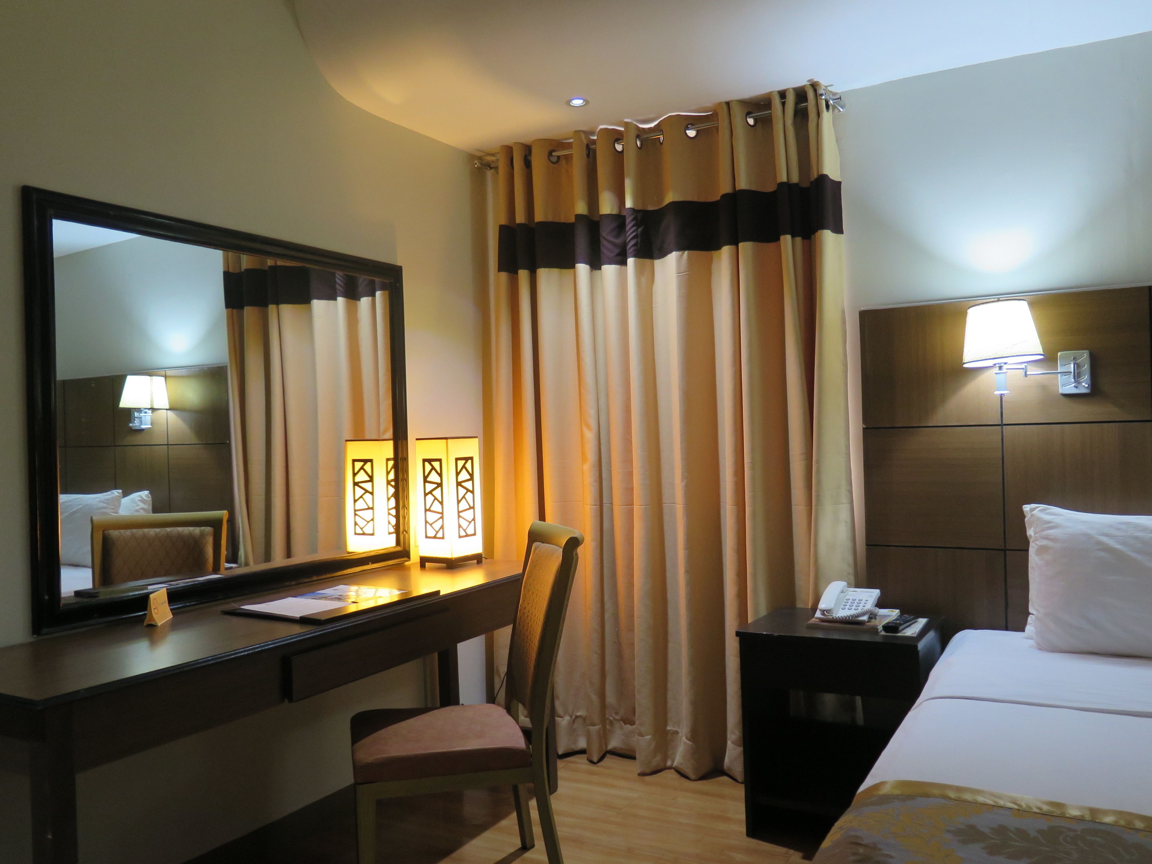 Ein Hotelzimmer mit einem Bett, einem Schreibtisch mit Stuhl, einem Nachttisch mit Lampe, einem Spiegel und einem Fenster mit Vorhängen.
