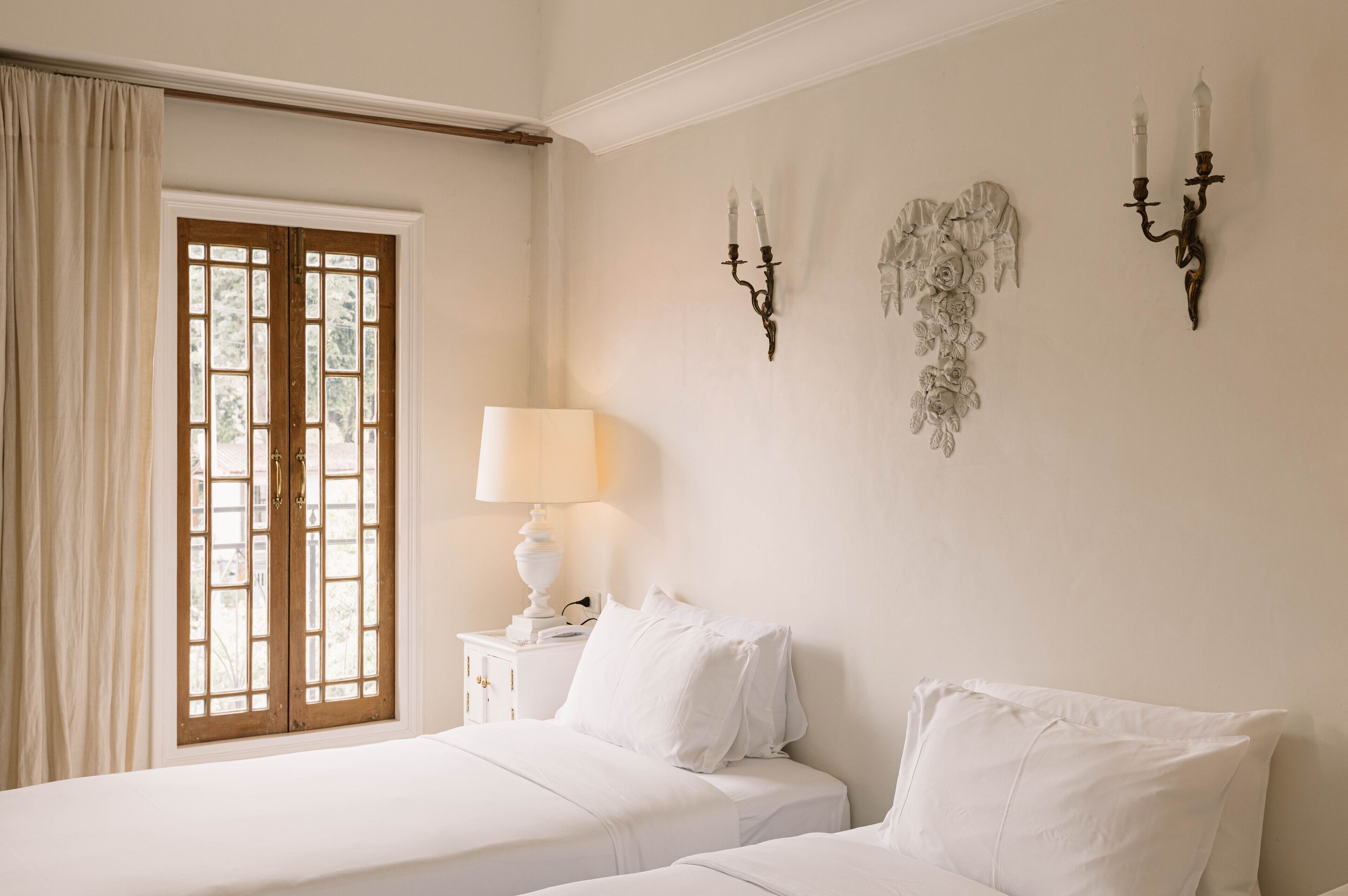 A hotel room with two beds, a window with curtains, a bedside table with a lamp, and wall-mounted candelabras.