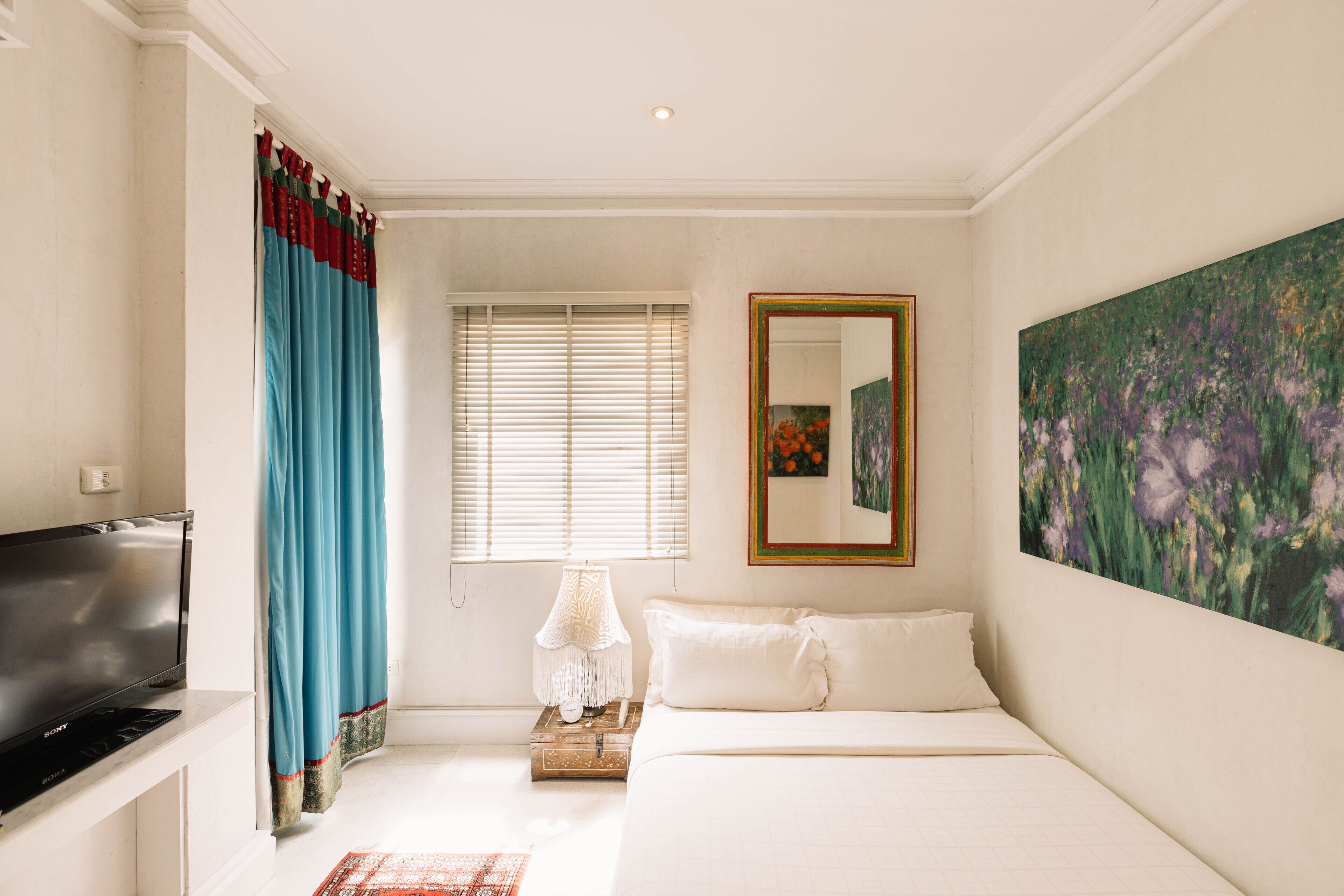 A bedroom with a bed, a TV, a window with blinds, a painting, and a mirror.