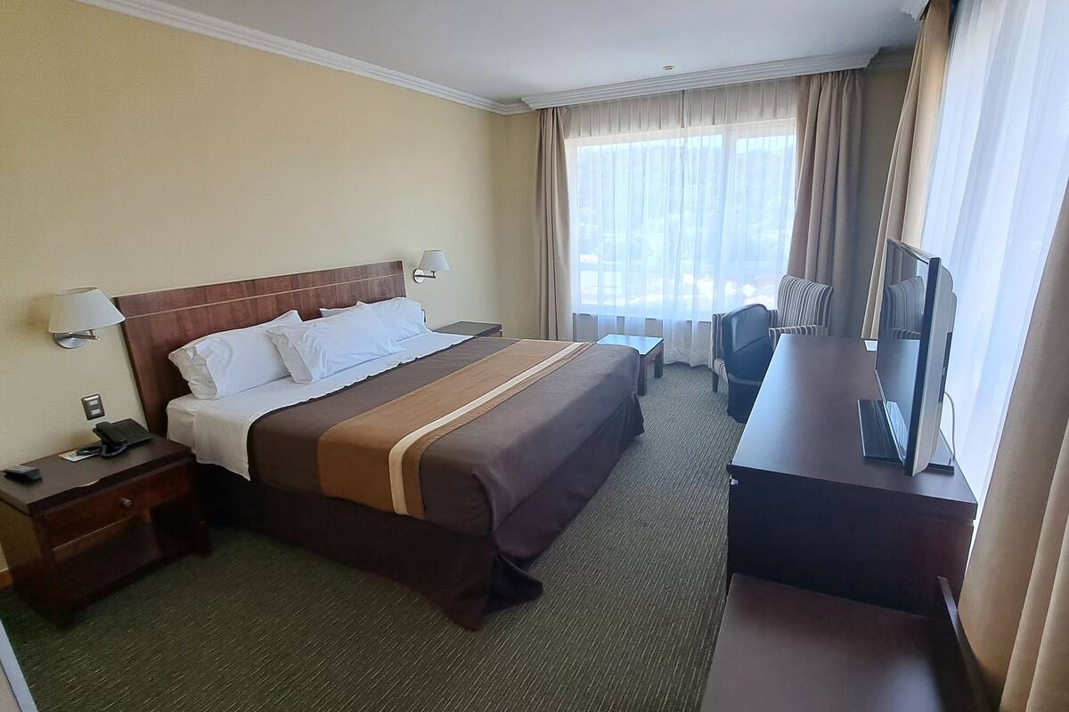 A hotel room with a bed, bedside tables, a desk with a chair, a TV, and a window with curtains.