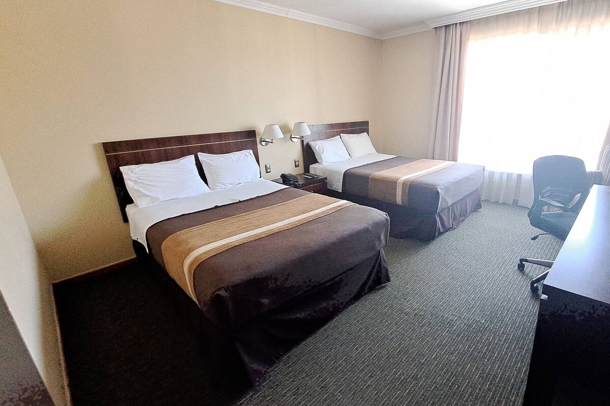 A hotel room with two beds, a desk, a chair, and a window with curtains.
