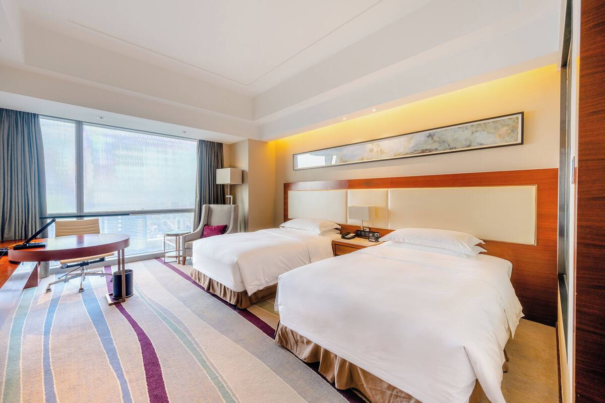 A hotel room with two beds, a desk, a chair, a lamp, and a large window.