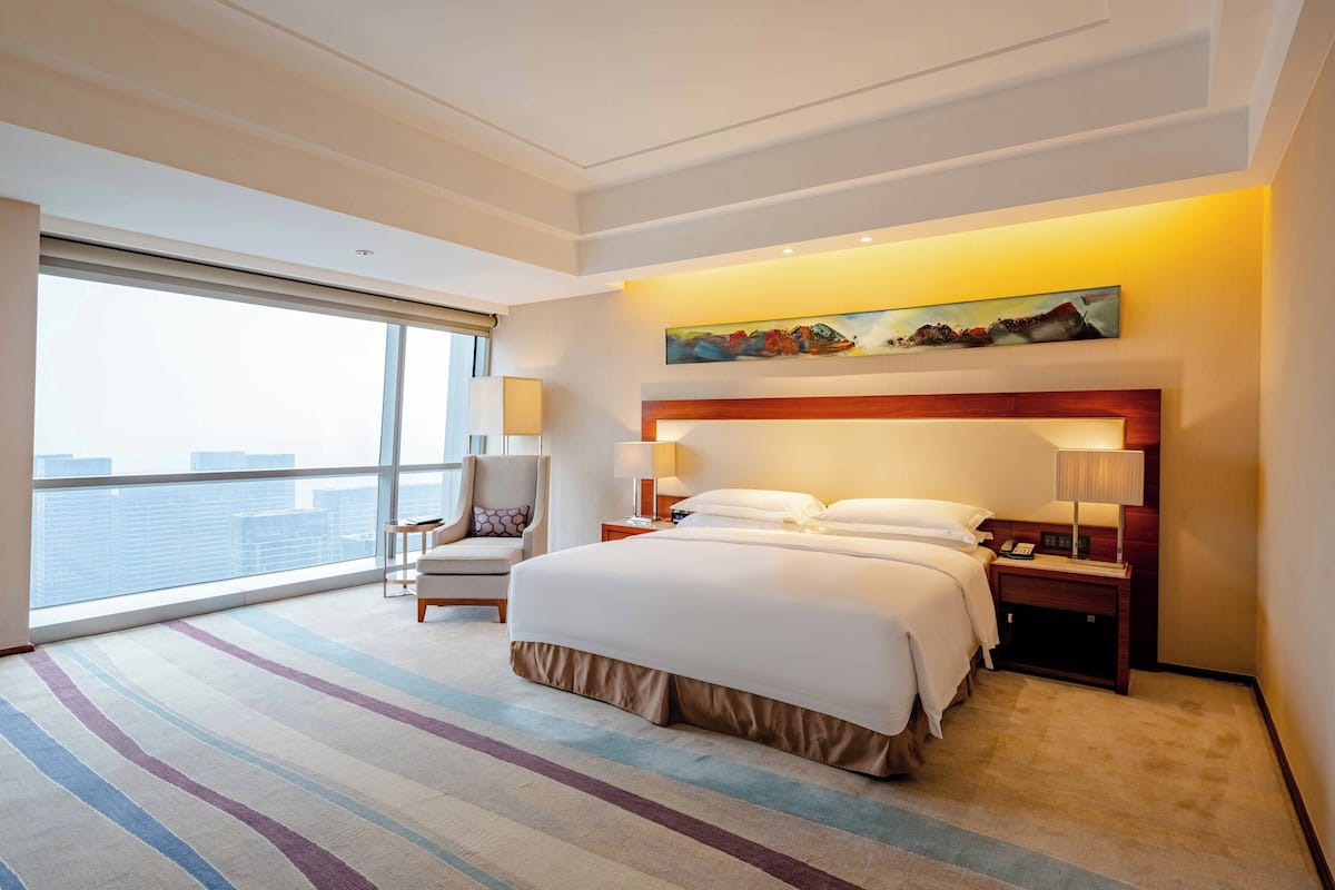A hotel room with a large bed, a nightstand with a lamp, a chair, and a large window with a view of the city.