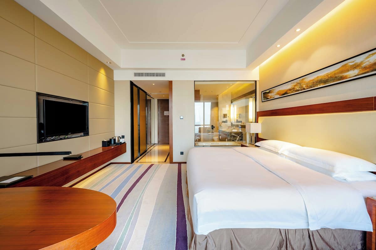 A hotel room with a large bed, a flat-screen TV, a wooden side table, and a striped area rug.