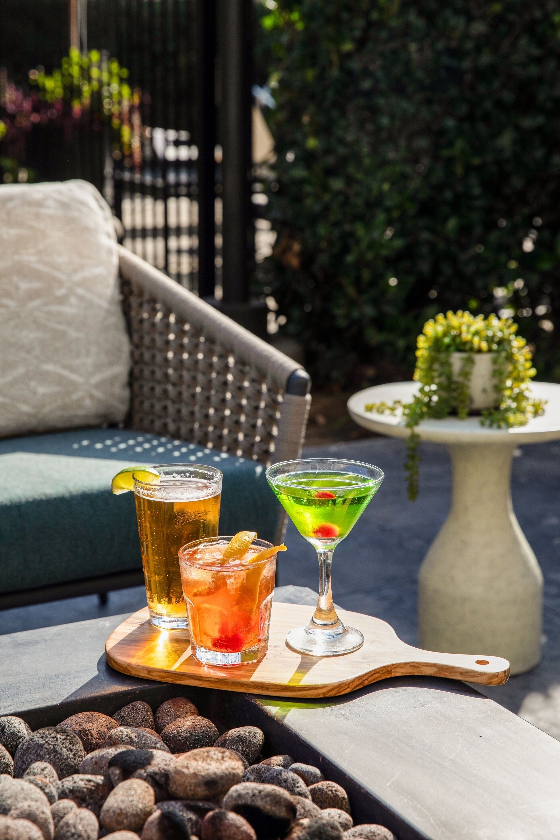 A table with two drinks and a martini glass, set on a wooden tray with pebbles.