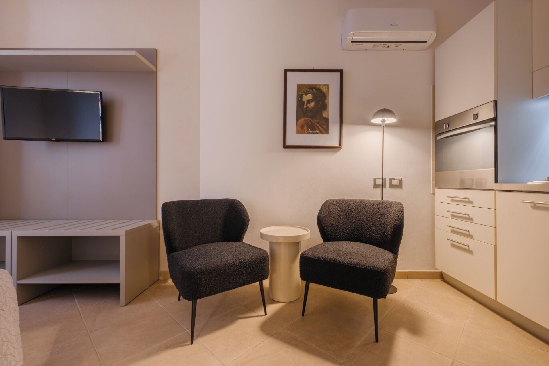Un salon moderne avec deux fauteuils sombres, une petite table ronde, un téléviseur à écran plat fixé au mur, une œuvre d’art encadrée au-dessus de la table, et un coin cuisine avec des placards et une cuisinière.
