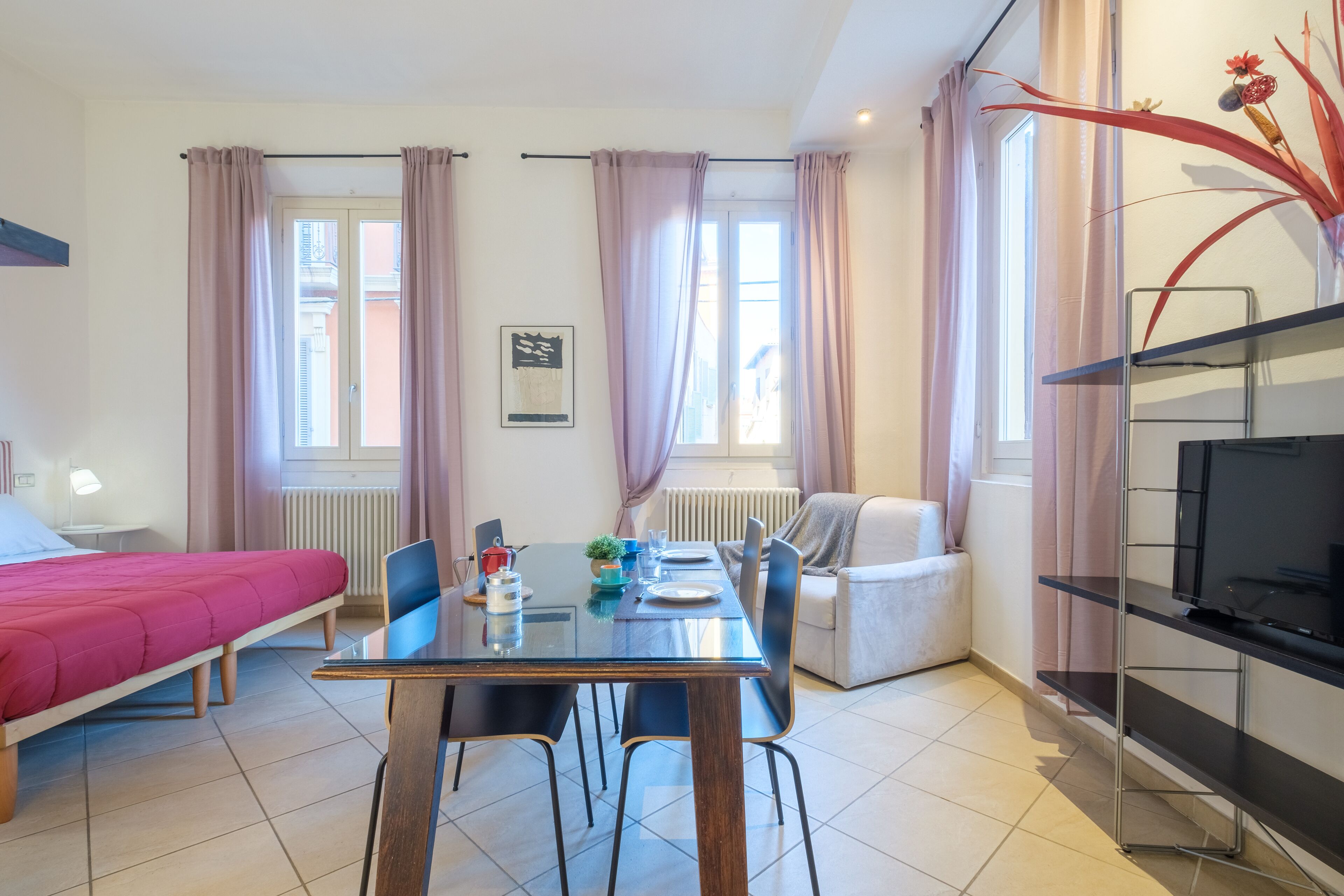 Ein modernes Hotelzimmer mit einem Bett, einem Esstisch mit Stühlen, einer Couch, einem Fernseher auf einem Schrank und großen Fenstern mit Vorhängen.