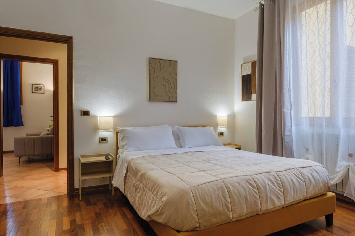 A bedroom with a bed, bedside tables, a wall-mounted artwork, and a view of a blue curtain and a chair in the adjacent room.