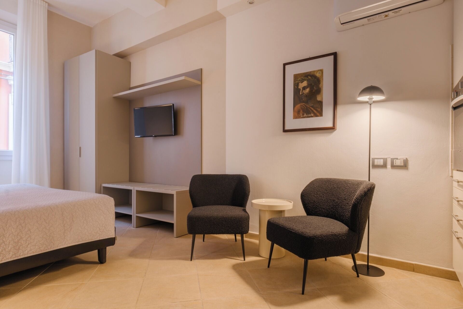 A modern hotel room with a bed, two armchairs, a desk, a flat-screen TV, a framed picture, a floor lamp, and an air conditioning unit.
