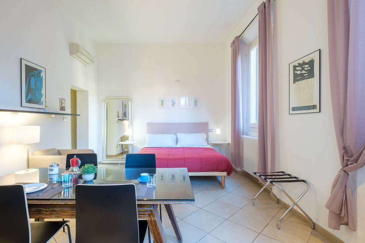 A hotel room with a bed, a dining table with chairs, a small kitchen area, and a bathroom visible through an open door.