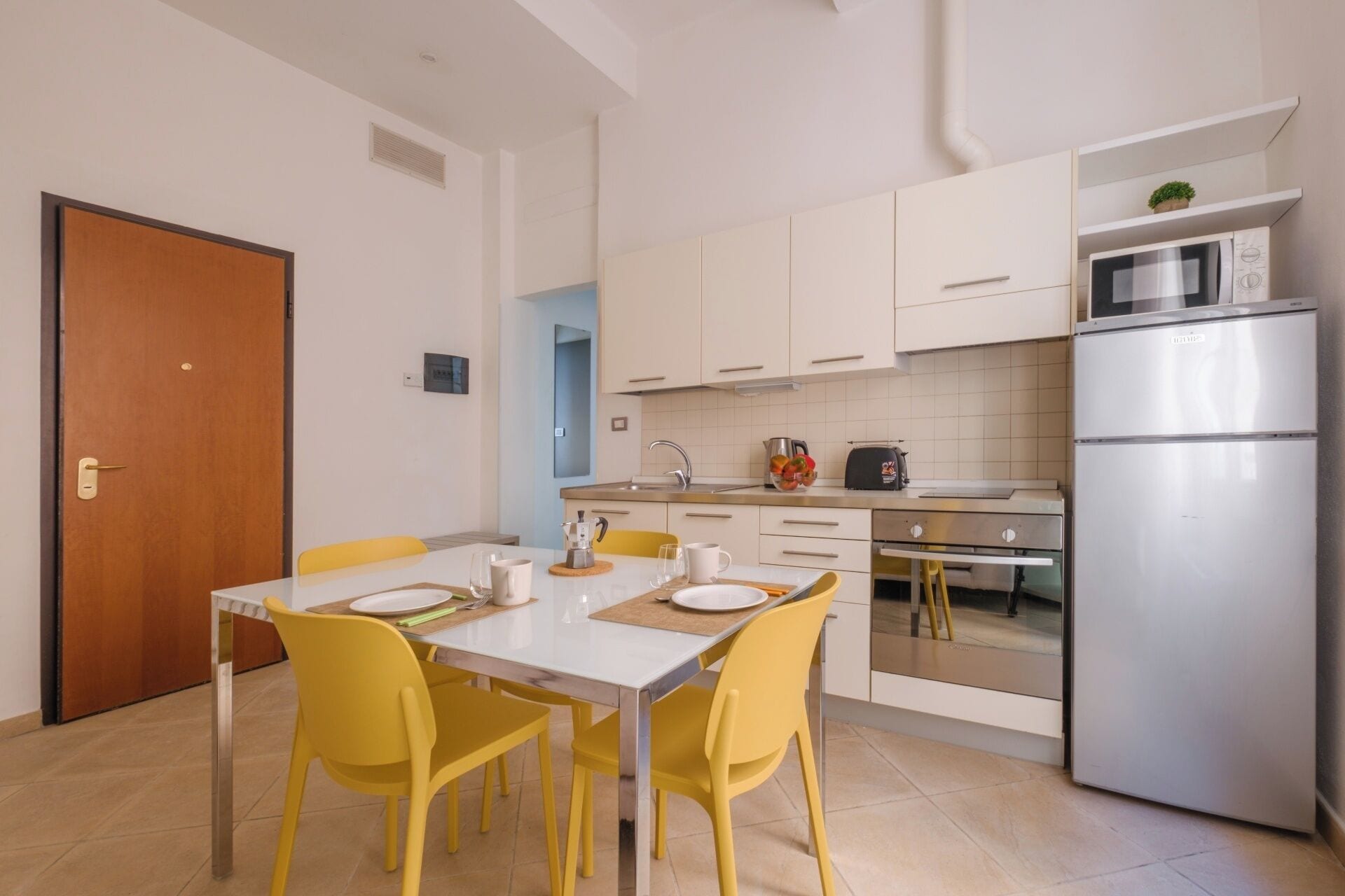 Une cuisine moderne avec des meubles blancs, un réfrigérateur en acier inoxydable et un coin repas avec des chaises jaunes et une table blanche.