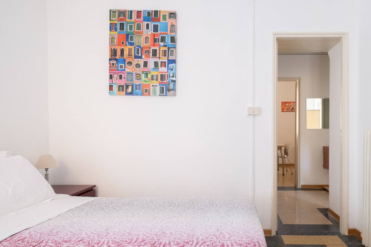 A hotel room with a bed, a nightstand, a lamp, and a colorful artwork on the wall.