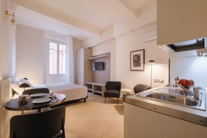 A compact hotel room with a bed, a small dining table with chairs, a kitchenette, a TV mounted on the wall, and a window with curtains. - Residenza Ariosto (Bologna)