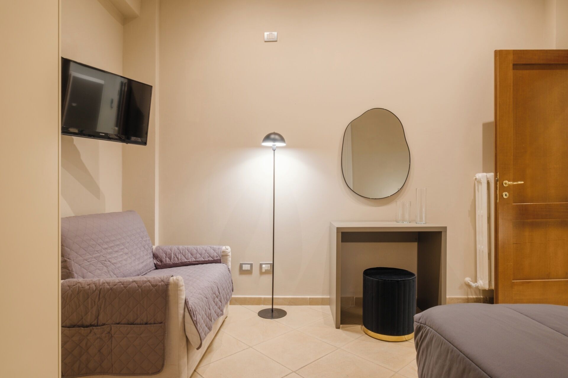 Un dormitorio con una cama, una silla, una lámpara de pie, un espejo, una chimenea y un televisor montado en la pared.